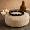 Bracelet tibétain en hématite noire posé sur un coussin de méditation beige, avec des bougies allumées et une statue de Bouddha floutée en arrière-plan, créant une ambiance zen et apaisante.