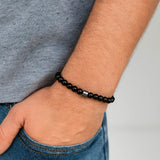 Tibetan Hematite Bracelet composé de perles noires polies avec une perle centrale en hématite, porté au poignet d’un homme en jean et t-shirt.