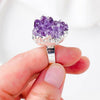 A ring with a silver band topped with a natural purple amethyst held in a hand.